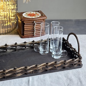 Könnte beinhalten: Ein rechteckiges Serviertablett mit einer dunkelgrauen Oberfläche und gewebten braunen Rändern. Drei klare Schnapsgläser mit Griffen stehen auf dem Tablett. Ein Holzuntersetzer-Set und eine dekorative Glasvase befinden sich im Hintergrund.