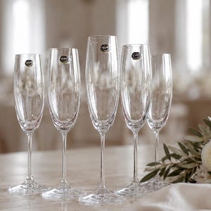Peut inclure: Un ensemble de cinq flûtes à champagne en verre transparent avec des pieds fins et des bases circulaires. Chaque flûte présente un petit autocollant circulaire près du bord. Les verres sont alignés sur une surface claire près de fleurs blanches.