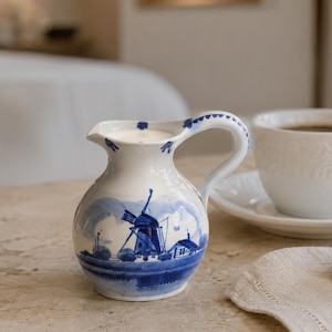 Könnte beinhalten: Ein weißes Milchkännchen aus Keramik mit einem blau handbemalten Windmühlen-Landschaftsmotiv, begleitet von einer weißen Teetasse auf passender Untertasse und einem goldbraunen Croissant auf einer hellen Stoffserviette.