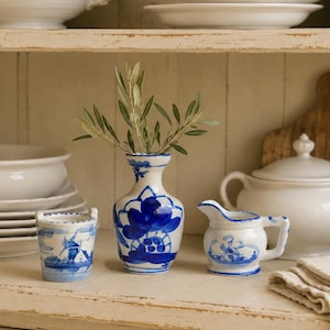 Puede incluir: Una pequeña colección de cerámica que incluye un jarrón pintado a mano en azul y blanco con motivos florales, un cubo en miniatura con una ilustración de molino de viento y una pequeña jarra decorada con una escena de molino, sobre un estante.