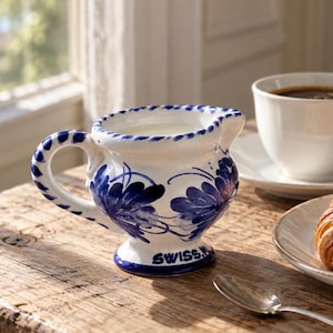 Peut inclure: Un pot à lait en céramique blanche orné de motifs floraux bleus peints à la main, avec une anse torsadée et une base sur pied. Le mot SWISS est inscrit en bleu sur la base. Le pot contient du lait et est placé près d'une tasse et d'un croissant.