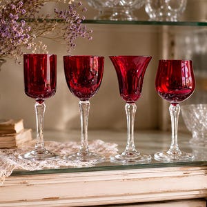 Peut inclure: Quatre verres à vin rouge rubis avec des tiges transparentes sont exposés sur une étagère en verre. Les verres ont des formes de cuvette différentes. L'étagère fait partie d'une armoire blanche avec une nappe en dentelle et un vase argenté avec des fleurs.