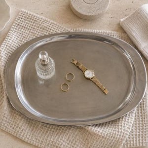 Pode incluir: Uma bandeja oval de metal prateado contendo um frasco de perfume de vidro transparente com tampa redonda, dois anéis simples de metal dourado e um relógio de pulso de metal dourado com pulseira de elos e mostrador branco.