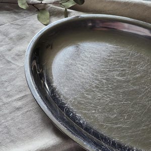 May include: An oval-shaped silver-toned metal tray with a polished finish. The surface of the tray displays numerous fine, visible surface scratches throughout the interior basin, while the outer rim remains smooth and reflective.