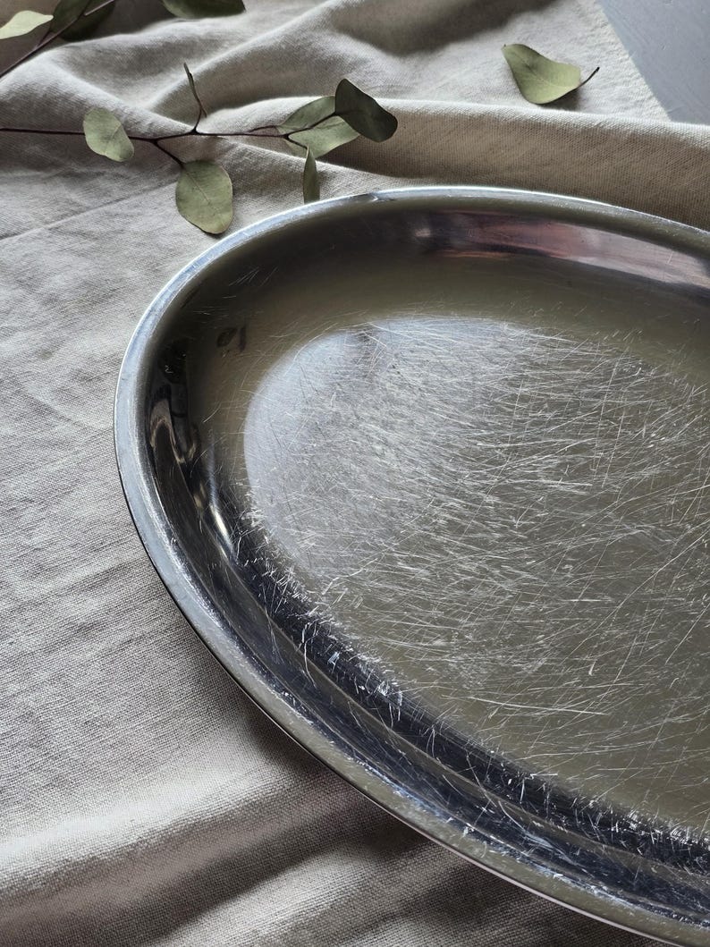 May include: An oval-shaped silver-toned metal tray with a polished finish. The surface of the tray displays numerous fine, visible surface scratches throughout the interior basin, while the outer rim remains smooth and reflective.