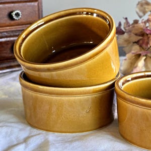 Può includere: Tre piccoli ramequin rotondi in ceramica con una smaltatura marrone dorato lucido sono impilati. I ramequin hanno un bordo leggermente svasato e sono su una superficie bianca. Un mobile in legno e fiori secchi sullo sfondo.