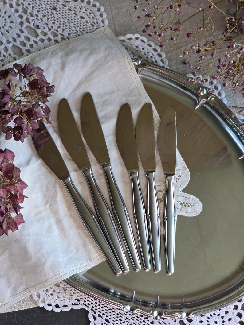 Pode incluir: Seis facas de manteiga de a&ccedil;o inoxid&aacute;vel dispostas sobre uma toalha de linho branca com acabamento em renda. As facas est&atilde;o em uma bandeja de servir prateada, com flores roxas secas ao fundo. Uma configura&ccedil;&atilde;o de mesa vintage.