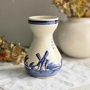 May include: Cream ceramic vase featuring a blue windmill design. The vase has a flared top with two blue stripes and a bulbous body. The windmill scene includes a windmill, trees, and houses, all in blue. The vase is displayed on a white cloth.