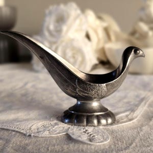 May include: A silver-toned, bird-shaped decorative dish. The dish has a long, curved spout and a rounded base. The bird's body is detailed with etched feather patterns. It sits on a white, lace-trimmed surface.