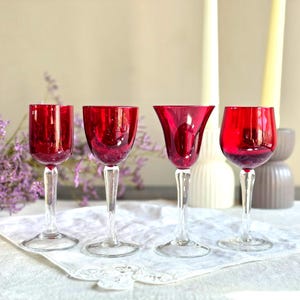May include: Four red wine glasses with clear stems and bases are arranged on a white lace cloth. The glasses have different bowl shapes. Purple flowers and candles are in the background.