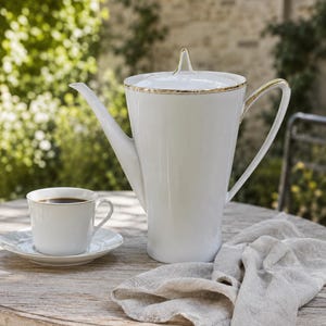 Könnte beinhalten: Eine hohe Kaffeekanne aus weißem Porzellan mit passendem Deckel und goldfarbenem Rand, begleitet von einer kleinen weißen Kaffeetasse und Untertasse, die jeweils mit einem dünnen Goldstreifen an den Rändern verziert sind.