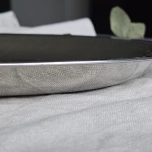 May include: A shallow, circular silver-toned metal tray with a smooth, reflective surface and a slightly raised rim. The tray features a polished metallic finish that catches the light, showcasing a clean, minimalist design with no additional patterns.