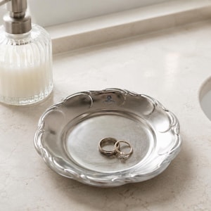 Op de afbeelding: Een ronde zilverkleurige metalen schaal met een geschulpte rand en een glad, reflecterend oppervlak. Twee ringen, een eenvoudige band en een ring met een kleine steen, liggen in het midden van de schaal. Een klein blauw logo is zichtbaar op de rand.