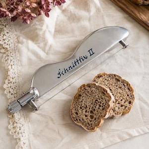 Pode incluir: Um cortador de pão de metal prateado com uma lâmina de corte de fio fino e um botão de ajuste de tensão. A lateral da ferramenta apresenta o texto azul "Schnittfix II" impresso em letra cursiva. Duas fatias de pão integral estão posicionadas por perto.