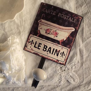 May include: A distressed metal sign with the words "CARTE POSTALE" and "LE BAIN". The sign depicts a bathtub with floral details. It's mounted on a black metal rod with a white oval knob. The sign has a vintage, rustic aesthetic.