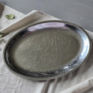 May include: An oval-shaped silver-toned metal serving platter with a polished, reflective surface. The platter features a smooth, slightly raised rim and shows visible fine surface scratches across the center area of the metal base.