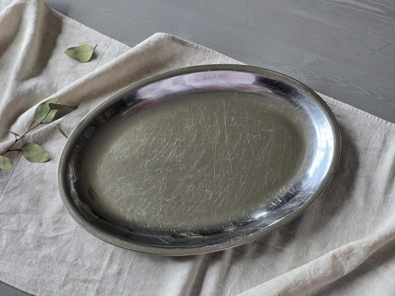 May include: An oval-shaped silver-toned metal serving platter with a polished, reflective surface. The platter features a smooth, slightly raised rim and shows visible fine surface scratches across the center area of the metal base.