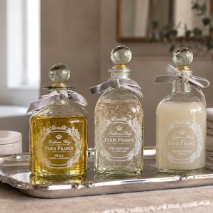 Op de afbeelding: Drie glazen flessen met bolvormige stoppen en grijze linten, elk voorzien van een wit etiket met de tekst "Paris France" en een kroonicoon. De flessen bevatten gele olie, heldere gel en witte shampoo, geplaatst op een zilveren dienblad.