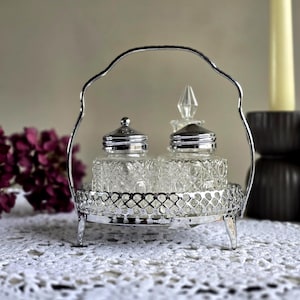 May include: A vintage silver-plated condiment set with two clear glass jars with ornate lids. The jars sit in a decorative silver tray with a handle. The set is on a white lace tablecloth, with purple flowers in the background.