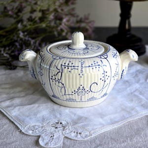 May include: A white ceramic sugar bowl with blue floral and linear patterns, featuring a lid, two handles, and a knob. The bowl rests on a white, lace-edged cloth, evoking a vintage or antique style. The measurements are in centimeters.