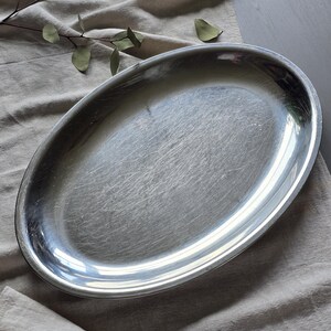 May include: An oval-shaped silver-colored metal serving platter with a smooth, reflective surface. The platter features a slightly raised rim and shows fine, visible surface scratches across its metallic finish, indicating regular use.