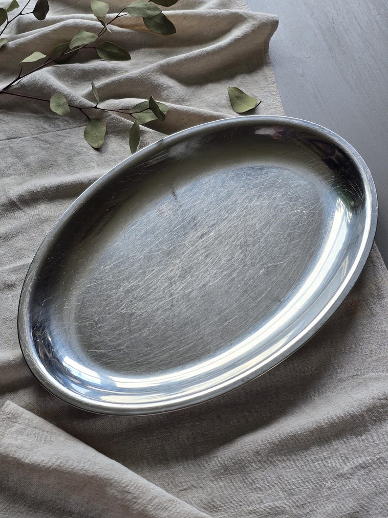 May include: An oval-shaped silver-colored metal serving platter with a smooth, reflective surface. The platter features a slightly raised rim and shows fine, visible surface scratches across its metallic finish, indicating regular use.