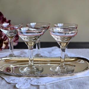 May include: Five clear champagne coupe glasses with a red rim, presented on a silver oval tray. The glasses have a stemmed design. The tray is placed on a white lace cloth, ideal for serving cocktails.