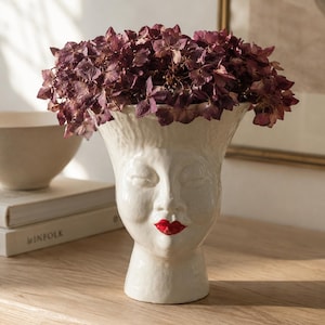 May include: A white ceramic head-shaped vase with a textured surface, featuring closed eyes and painted red lips. The vase is filled with a cluster of dried, deep purple hydrangea flowers, creating a contrast between the white ceramic and the dark petals.