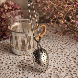 Peut inclure: Un infuseur à thé en métal argenté doté d'une longue poignée en forme de ciseaux et d'un panier perforé en forme d'œuf. L'infuseur est placé à la verticale à côté d'une tasse en verre transparent dans un support en métal argenté ajouré.