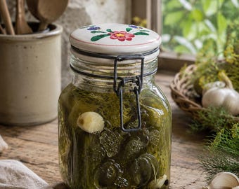 May include: A clear glass jar filled with green pickles, garlic cloves, and spices. The jar features embossed fruit designs on the sides and a metal wire bail closure. It is topped with a white ceramic lid decorated with a hand-painted floral motif.