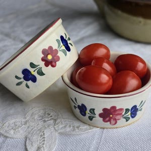 Op de afbeelding: Twee kleine, crèmekleurige keramische kommen met bloemmotieven. Eén kom is gevuld met rode tomaten. De kommen hebben een rode rand en zijn versierd met rode, blauwe en groene bloemen. De kommen staan op een wit, getextureerd oppervlak.