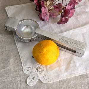 May include: A silver citrus juicer with the text "THE ORIGINAL PEACHTREE" next to a yellow lemon on a white doily. Dried pink flowers are in the background. The juicer is designed to extract juice from citrus fruits.
