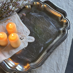 Op de afbeelding: Een vintage zilverkleurige dienblad met een geschulpte rand. Drie brandende, oranje kaarsen liggen op een wit servet met kanten rand, naast een takje paarse bloemen. De dienblad staat op een neutraal gekleurd linnen tafelkleed.