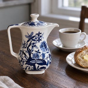 May include: A white ceramic coffee pot featuring a detailed blue floral pattern, a matching lid with a knob handle, a curved spout, and a side handle. The intricate design depicts various flowers and foliage wrapping around the body of the pot.