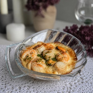 Può includere: Un piatto di vetro trasparente pieno di frutti di mare, tra cui gamberi, capesante e cozze, in una salsa cremosa. Il piatto è guarnito con erbe verdi e ha una crosta dorata. Il piatto è su una tovaglia di pizzo bianco.