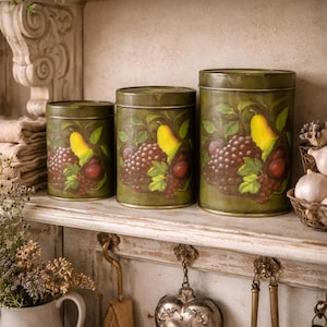 Può includere: Tre barattoli di metallo verde oliva con coperchi, ognuno decorato con un disegno dipinto di pere gialle, uva rossa e foglie verdi. I barattoli sono disposti su una mensola bianca invecchiata, evocando un'estetica da cucina vintage.