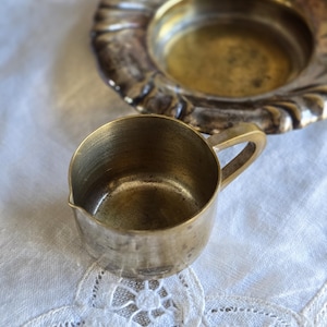 Op de afbeelding: Een kleine, antieke messing roomkannetje met een handvat en tuit, naast een decoratieve messing schaal. Beide items hebben een aangeslagen, verouderde uitstraling, wat suggereert dat ze vintage zijn. Ze liggen op een witte, met kant afgezette doek.