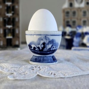 Może przedstawiać: Białe jajko w niebiesko-białym porcelanowym kieliszku do jajek. Kieliszek do jajek ma wzór sceny morskiej. Kieliszek do jajek stoi na białej serwetce z koronkową krawędzią. Ozdobne budynki w tle.