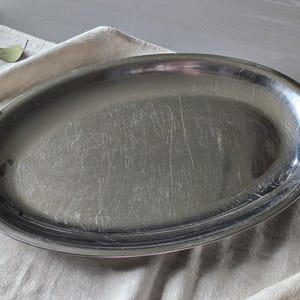 May include: An oval-shaped silver-toned metal serving platter with a smooth, reflective surface. The platter features a raised rim and shows visible surface scratches across the center, indicating previous use and a metallic finish.