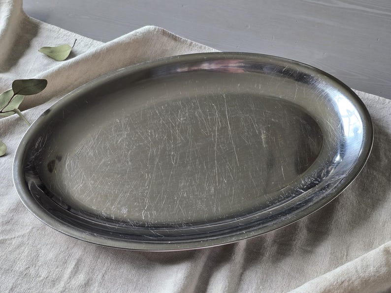 May include: An oval-shaped silver-toned metal serving platter with a smooth, reflective surface. The platter features a raised rim and shows visible surface scratches across the center, indicating previous use and a metallic finish.