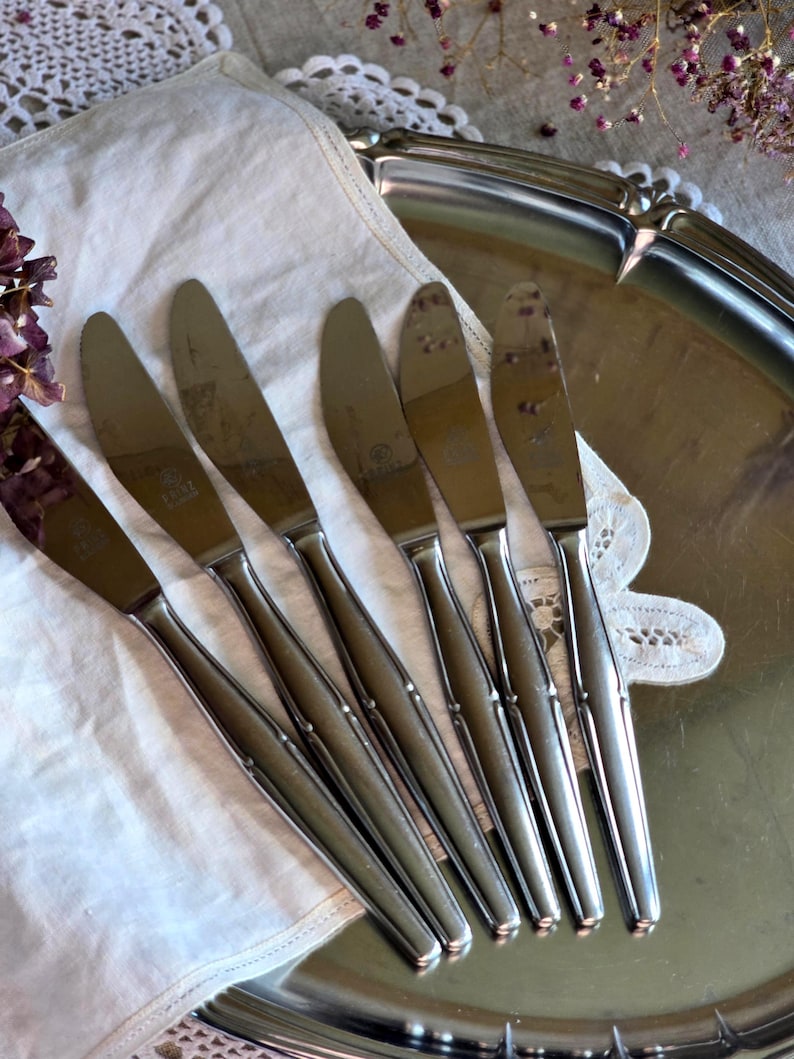 Pode incluir: Um conjunto de seis facas de manteiga de a&ccedil;o inoxid&aacute;vel dispostas sobre uma toalha de linho branca, colocadas em uma bandeja de servir prateada. As facas t&ecirc;m um design simples e elegante com uma l&acirc;mina ligeiramente curvada e uma al&ccedil;a fina. O fundo inclui renda e flores secas.