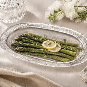 May include: An oval, textured glass serving dish filled with green asparagus spears and garnished with lemon slices. The dish sits on a neutral-colored tablecloth, with a decorative glass bowl and white flowers in the background.