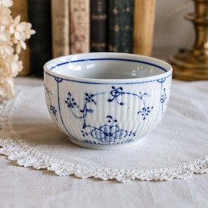Może przedstawiać: Biała ceramiczna miska z niebieskim kwiatowym wzorem. Miska ma żebrowaną fakturę i falisty brzeg. Niebieski wzór przedstawia motywy kwiatowe i winorośli. Miska stoi na białej koronkowej serwetce.