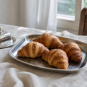 May include: A rectangular stainless steel tray holding four golden-brown, flaky croissants, positioned next to a small, polished silver-toned butter dish with a lid. The items are arranged on a textured, light-coloured fabric surface.