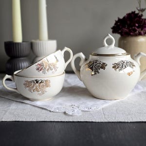 May include: A cream-coloured vintage tea set with gold floral patterns. The set includes a lidded sugar bowl and two teacups. The teacups are stacked, and the set is arranged on a white doily. A vase of flowers and candles are in the background.