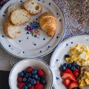 Peut inclure: Deux assiettes rondes en céramique et un bol assorti avec une base blanche, des bords bleus et un motif floral délicat. Les assiettes contiennent des tranches de baguette, un croissant, des œufs brouillés, du pain grillé, des fraises et des myrtilles.