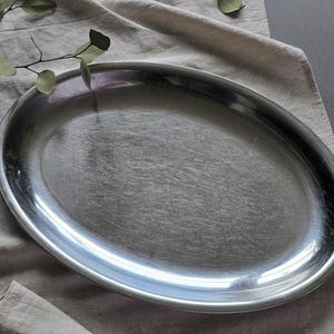 May include: An oval-shaped serving platter crafted from polished stainless steel with a smooth, reflective surface and a slightly raised rim. The platter features a simple, clean design with a metallic silver finish that catches the light.