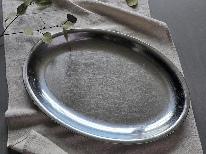 May include: An oval-shaped serving platter crafted from polished stainless steel with a smooth, reflective surface and a slightly raised rim. The platter features a simple, clean design with a metallic silver finish that catches the light.