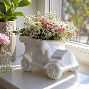 Könnte beinhalten: Ein weißer Keramikübertopf in Form eines Oldtimers, gefüllt mit einem bunten Blumenstrauß aus weißen Gänseblümchen, rosa Nelken und kleinen weißen Schleierkrautblüten. Der Übertopf hat eine glänzende Oberfläche und detaillierte Räder.
