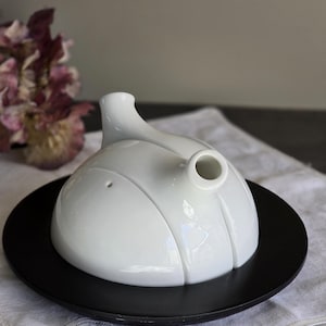 May include: A white ceramic teapot with a rounded body and a spout, resting on a black plate. The teapot has a unique design with vertical grooves. The plate is circular and has a matte finish. The background includes a white cloth and a blurred floral arrangement.